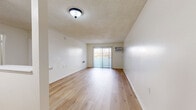 Located on Wyandotte Street in the Reseda neighborhood of Los Angeles, this one-bedroom, one-bathroom unit offers a well-organized single-floor layout with consistent light wood flooring throughout. The living room is a spacious, open area highlighted by a sliding glass door that leads to a balcony, allowing natural light to fill the room. A wall-mounted air conditioning unit provides climate control, and a textured ceiling adds a classic residential character. The kitchen is positioned adjacent to the living area, featuring dark wood cabinetry, a built-in countertop with a pass-through bar, and a refrigerator, creating a practical and accessible cooking space. A dedicated closet is conveniently accessible from the main living area. The bedroom offers a comfortable amount of space with a window for natural light and a mirrored sliding closet with gold-tone frames, contributing both storage and a sense of added depth to the room. The bathroom is clean and functional, equipped with light wood-style flooring, a combined tub and shower unit with safety grab bars, a handheld showerhead, and a vanity. Throughout the unit, neutral white walls and light flooring create a bright, cohesive aesthetic.
