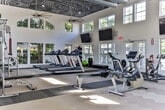 Located at 1000 Shadowood Pkwy SE in Atlanta, Georgia, this well-appointed fitness center offers a spacious, single-floor layout designed to support a full range of workout routines. The facility is defined by its soaring ceilings, with arched clerestory windows and multiple large windows along the walls that flood the interior with natural light and offer views of the surrounding greenery.

The cardio zone is generously equipped with treadmills, elliptical machines, stationary bikes, and a rowing machine, all arranged to allow comfortable spacing between users. A dedicated strength training area features a multi-station cable machine, adjustable benches, and a rack of free weights, supported by wall-mounted mirrors that enhance the sense of openness. A separate functional training nook houses stability balls, kettlebells, resistance tools, and a dumbbell rack, making it ideal for dynamic or accessory workouts.

Decorative ceiling details add visual character to portions of the space, while exposed industrial ductwork lends an energetic, modern atmosphere. Wall-mounted flat-screen televisions are positioned throughout for entertainment during workouts. French doors open directly to an outdoor area, providing easy access and additional ventilation. A water cooler station ensures hydration is readily available. The combination of thoughtful design, ample equipment, and bright, airy surroundings makes this fitness center a well-rounded and inviting facility.