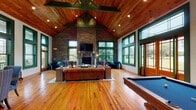 Located at 1000 Flycatcher Way in Arden, North Carolina, this single-floor gathering space showcases a masterful blend of rustic craftsmanship and inviting comfort. The centerpiece of the main living area is a dramatic floor-to-ceiling stacked stone fireplace wall, complete with a mounted flat-screen TV and a stone mantel, creating a striking focal point that anchors the room. Soaring vaulted ceilings clad in warm, rich cedar planks rise overhead, supported by bold exposed timber trusses finished in deep green, lending an authentic lodge-style character throughout. Gleaming natural hardwood floors extend across the entire space, adding warmth underfoot. Generous windows trimmed in forest green line the walls, allowing abundant natural light to pour in while framing views of the surrounding lush greenery. The open layout accommodates distinct areas, including a comfortable lounge seating arrangement with leather and velvet upholstered furnishings gathered around the fireplace, a dedicated pool table area ideal for recreation, and a dining space featuring a rustic wood table and buffet sideboard. Shiplap white walls contrast beautifully with the stone and wood elements throughout. The overall ambiance is warm, welcoming, and distinctly Appalachian in character, making this a well-appointed communal retreat.