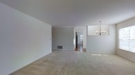 This single-floor space presents a straightforward, open layout with a clean, neutral aesthetic throughout. The main living area is finished with wall-to-wall carpeting in a warm beige tone, offering a soft and comfortable underfoot feel. The walls are painted in a light grey-white hue, contributing to a bright, airy atmosphere that feels open and versatile.

A notable design element is the elegant multi-arm chandelier suspended from the ceiling in the adjacent dining or transitional area, adding a traditional decorative touch to the otherwise minimalist space. The ceiling is smooth and white, enhancing the sense of openness.

A hallway visible from the main living area leads toward what appears to be a kitchen, where hardwood-style flooring transitions from the carpeted areas, and natural light filters through a window, illuminating the space. A floor vent is also visible, indicating a central forced-air system.

The open sightlines between the living, dining, and kitchen areas create a cohesive, connected flow throughout the single level, making the space feel functional and easy to navigate. The overall ambiance is neutral, move-in ready, and well-suited for a range of furnishing styles.