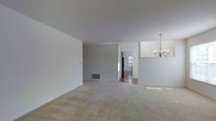 This single-floor space presents a straightforward, open layout with a clean, neutral aesthetic throughout. The main living area is finished with wall-to-wall carpeting in a warm beige tone, offering a soft and comfortable underfoot feel. The walls are painted in a light grey-white hue, contributing to a bright, airy atmosphere that feels open and versatile.

A notable design element is the elegant multi-arm chandelier suspended from the ceiling in the adjacent dining or transitional area, adding a traditional decorative touch to the otherwise minimalist space. The ceiling is smooth and white, enhancing the sense of openness.

A hallway visible from the main living area leads toward what appears to be a kitchen, where hardwood-style flooring transitions from the carpeted areas, and natural light filters through a window, illuminating the space. A floor vent is also visible, indicating a central forced-air system.

The open sightlines between the living, dining, and kitchen areas create a cohesive, connected flow throughout the single level, making the space feel functional and easy to navigate. The overall ambiance is neutral, move-in ready, and well-suited for a range of furnishing styles.