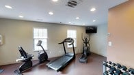 This single-floor fitness room offers a dedicated space for cardiovascular exercise, outfitted with a curated selection of cardio equipment including a treadmill, stationary bike, and elliptical trainer. The room features a durable, dark rubberized flooring that provides both safety and functionality underfoot, ideal for high-traffic workout use.

Natural light filters into the space through multiple windows fitted with white horizontal blinds, creating a bright and comfortable environment for exercise. Recessed lighting in the white ceiling supplements the natural light, ensuring the room remains well-lit at any time of day. A wall-mounted flat-screen television is positioned at an optimal viewing angle, allowing users to stay entertained or follow workout programming during their sessions.

The neutral-toned walls and clean, minimalist design keep the focus on functionality. The ceiling-mounted HVAC vent ensures proper climate control, contributing to a comfortable workout environment year-round. The overall layout is efficient and purposeful, making the most of the available space while keeping equipment accessible and easy to navigate. This fitness room provides a practical and convenient option for those looking to maintain an active lifestyle without leaving the premises.