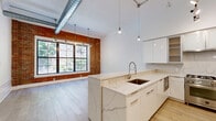 Located at 100 Paterson Plank Rd in Jersey City, NJ, this thoughtfully designed one-bedroom, one-bathroom residence blends industrial character with modern refinement across a generously sized single floor. The open living area immediately impresses with soaring ceilings, exposed brick accent walls, and large steel-framed grid windows that flood the space with natural light and frame vibrant green outdoor views. Light oak hardwood flooring runs throughout, tying the rooms together seamlessly. The chef's kitchen is a standout feature, boasting sleek high-gloss white cabinetry, marble-look stone countertops and backsplash, stainless steel appliances including a professional-grade gas range, and pendant lighting suspended from the dramatic ceiling height — creating an elegant, functional workspace. The spacious primary bedroom continues the bright, airy aesthetic with oversized windows and clean white walls. A dedicated walk-in closet offers extensive built-in shelving and hanging rods, providing exceptional storage. The bathroom is finished to a high standard with dark large-format floor tiles, a wood-toned floating vanity, a full-width mirror, and a glass-enclosed shower stall. The overall ambiance is one of urban sophistication, balancing loft-style industrial elements with refined contemporary finishes.