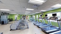 This fitness center is housed on a single level and designed to accommodate a range of cardio-focused workouts. The space is characterized by its vibrant green accent walls, which contrast with the light-toned luxury vinyl flooring and create an energizing environment. The suspended drop ceiling features a grid of recessed fluorescent panel lights, ensuring consistent, bright illumination throughout the room.

The cardio equipment area is well-stocked with multiple treadmills and elliptical machines arranged in organized rows, providing ample capacity for simultaneous users. Personal viewing screens are mounted on select machines, and wall-mounted flat-screen televisions are positioned for visibility across the floor. Large pedestal fans are strategically placed along the walls to maintain airflow and comfort during workouts.

Mirrored wall sections reflect light and enhance the perceived spaciousness of the room. Windows along one wall allow natural light to filter in, adding to the open feel. A designated stretching or free-weight area is visible toward one end of the space. Overall, the fitness center maintains a clean, functional, and motivating atmosphere well-suited for daily exercise routines.