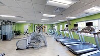 This fitness center is housed on a single level and designed to accommodate a range of cardio-focused workouts. The space is characterized by its vibrant green accent walls, which contrast with the light-toned luxury vinyl flooring and create an energizing environment. The suspended drop ceiling features a grid of recessed fluorescent panel lights, ensuring consistent, bright illumination throughout the room.

The cardio equipment area is well-stocked with multiple treadmills and elliptical machines arranged in organized rows, providing ample capacity for simultaneous users. Personal viewing screens are mounted on select machines, and wall-mounted flat-screen televisions are positioned for visibility across the floor. Large pedestal fans are strategically placed along the walls to maintain airflow and comfort during workouts.

Mirrored wall sections reflect light and enhance the perceived spaciousness of the room. Windows along one wall allow natural light to filter in, adding to the open feel. A designated stretching or free-weight area is visible toward one end of the space. Overall, the fitness center maintains a clean, functional, and motivating atmosphere well-suited for daily exercise routines.