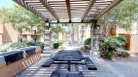 This outdoor communal space is defined by a sturdy pergola structure supported by stone masonry columns, casting a striking pattern of light and shadow across the area beneath. The pergola provides partial shade over a metal mesh picnic table with attached bench seating, making it a practical gathering spot for residents. Built-in grilling stations are positioned to one side, offering convenient outdoor cooking facilities. The surrounding landscape features a thoughtful mix of desert-adapted shrubs, ornamental grasses, and mature trees — including tall palms and leafy shade trees — that lend the space a lush, established feel. Pathway lighting is integrated into the design via globe-style lamp posts, supporting use of the area during evening hours. The hardscaped ground beneath the pergola transitions into a maintained pathway that winds through the greenery toward the residential buildings in the background, which feature a warm stucco exterior typical of Southwest architecture. The overall ambiance is relaxed and community-oriented, balancing functional amenities with natural surroundings in an inviting outdoor setting.