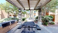This outdoor communal space is defined by a sturdy pergola structure supported by stone masonry columns, casting a striking pattern of light and shadow across the area beneath. The pergola provides partial shade over a metal mesh picnic table with attached bench seating, making it a practical gathering spot for residents. Built-in grilling stations are positioned to one side, offering convenient outdoor cooking facilities. The surrounding landscape features a thoughtful mix of desert-adapted shrubs, ornamental grasses, and mature trees — including tall palms and leafy shade trees — that lend the space a lush, established feel. Pathway lighting is integrated into the design via globe-style lamp posts, supporting use of the area during evening hours. The hardscaped ground beneath the pergola transitions into a maintained pathway that winds through the greenery toward the residential buildings in the background, which feature a warm stucco exterior typical of Southwest architecture. The overall ambiance is relaxed and community-oriented, balancing functional amenities with natural surroundings in an inviting outdoor setting.