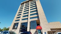 This prominent commercial building rises several stories above its urban surroundings, featuring a clean, modern facade clad in light-colored stone panels with a grid of large windows running across each floor. At street level, a distinctive Wells Fargo branch anchors the building's ground floor, marked by its recognizable red canopy entrance and floor-to-ceiling glass storefront that invites natural light into the interior. The building's bold, angular architectural design gives it a strong presence within the downtown streetscape.

The surrounding environment adds significant context to the space. A spacious public plaza nearby features broad paved brick walkways, open greenery, and a small auxiliary structure with decorative ironwork details, all set beneath open skies. This pedestrian-friendly area provides a welcoming outdoor environment with views of neighboring historic architecture, including a notable Victorian-style building with ornate detailing, as well as other mid-rise commercial and residential structures that characterize the urban core. The combination of the tower's contemporary commercial design and the open plaza creates a dynamic, accessible environment suited to both business and community activity.