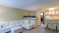 This single-floor space presents an open-concept layout that promotes a seamless flow between the living area and surrounding rooms. The living room is anchored by plush, light-colored carpeting and features warm, yellow-toned walls that lend a welcoming ambiance. A white tufted sectional sofa with a matching ottoman serves as the focal point of the space, positioned beneath a textured ceiling that adds subtle visual interest overhead.

A low half-wall divider separates the living area from an adjoining space, topped with decorative planters containing tall grass arrangements, providing a stylish yet functional partition. This architectural detail maintains an open feel while defining distinct zones within the layout. A ceiling fan is visible beyond the divider, suggesting a secondary living or dining area.

A hallway leads further into the unit, connecting to additional rooms, with a window at its end drawing in natural light. To the right, a glimpse of a counter and bar-style seating hints at a kitchen or dining space. The overall design is clean and neutral, with dark baseboards providing a grounding contrast to the lighter walls and flooring throughout.