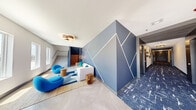 This thoughtfully designed lobby and hallway space makes a strong first impression through its striking architectural details and cohesive contemporary aesthetic. The centerpiece of the lounge area is a dramatic blue geometric accent wall adorned with angular LED light strips that create a dynamic, eye-catching focal point. The lounge is furnished with curved, vibrant blue chairs, a plush neutral-toned sofa accented with teal throw pillows, and natural wood side tables, all arranged atop a layered area rug that ties the color palette together. Large windows along the exterior wall flood the seating area with abundant natural light while offering views of the surrounding outdoors. Polished large-format tile flooring grounds the lounge, transitioning seamlessly to a richly patterned blue-and-white carpet that extends through a long, well-appointed corridor. The hallway features warm overhead lighting and dark wood-toned door surrounds that add warmth and definition to the space. A staircase with a sleek metal handrail and herringbone wood-paneled landing is visible from the lounge, adding an elegant architectural layer. The overall ambiance is modern, professional, and inviting, with a carefully curated color scheme of blue, grey, and white that lends the space a cohesive, upscale character throughout.