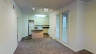This single-floor residence offers a practical and cohesive open-concept layout that seamlessly connects the kitchen and living areas. The kitchen is well-appointed with white cabinetry, dark countertops, and a full suite of white appliances, including a refrigerator, electric range, and over-the-range microwave. A curved center island provides additional counter space and serves as a natural dividing point between the kitchen and the adjacent living area. A wine rack adds a thoughtful detail above the cabinetry. Track lighting illuminates the kitchen area, while a flush-mount fixture adds additional brightness, keeping the space well-lit throughout. The living area is finished with wall-to-wall carpeting, lending a warm and comfortable feel underfoot. Crown molding lines the ceilings, adding a refined architectural touch throughout the space. A full-panel door with a tall sidelight window allows natural light to filter into the interior, enhancing the overall brightness of the open-plan layout. The combination of neutral tones, functional design, and clean finishes gives this residence a welcoming and move-in-ready atmosphere suited for everyday living.
