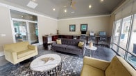 This single-floor business center offers a well-appointed, multifunctional space that seamlessly blends productivity with comfort. The room features vaulted ceilings fitted with a ceiling fan and recessed lighting, lending an open and airy feel throughout. Crown molding adds a refined architectural touch to the neutral-toned walls, while polished concrete floors provide a sleek, modern foundation.

The lounge area is anchored by a tufted charcoal sofa and gold accent armchairs arranged around a distinctive marble-top coffee table and a patterned area rug, creating an inviting atmosphere for relaxed collaboration or casual gatherings. A wall-mounted flat-screen television is positioned on an accent wall painted in a muted olive-green tone, adding a pop of color to the space.

Along one wall, a built-in granite-topped workstation with dark wood cabinetry accommodates multiple computer workstations with ergonomic chairs, providing ample space for focused work. Framed artwork in teal and green tones accents the walls above the workstations.

Large floor-to-ceiling windows and glass doors flood the space with natural light and offer direct views of an outdoor pool and patio area. A glass-door entryway connects to an adjacent corridor, ensuring easy accessibility throughout the facility.