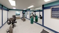Located at 500 Harry S Truman Drive in Largo, Maryland, this single-floor fitness center is designed to provide a functional and motivating environment for exercise. The space is immediately distinguished by its bold interior design, featuring a striking combination of teal and navy blue accent bands that run along the upper and lower portions of the white walls, lending the room a clean and energetic aesthetic.

The fitness area is equipped with a variety of cardio machines, including a treadmill, elliptical trainer, and recumbent exercise bike, offering users a well-rounded selection of cardiovascular workout options. A full-length mirror spans a large section of one wall, visually expanding the space and allowing users to monitor their form during exercise. Rolled yoga or exercise mats are stored in a wire basket, indicating availability of floor-based workouts as well.

The flooring consists of speckled, light-colored terrazzo-style material, which is both durable and easy to maintain — well-suited for a high-use fitness environment. Recessed fluorescent panel lighting illuminates the space evenly from above, while windows along one wall allow natural light to filter in and provide views of the surrounding outdoor greenery. Motivational framed artwork adorns the walls, reinforcing the space's active and encouraging atmosphere. A door with a glass panel provides access to an adjoining area.