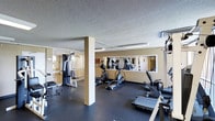 This fitness center at 2038 South Vaughn Way in Aurora, Colorado offers a well-equipped exercise space designed to support a range of workout routines. The single-level layout features a dark, cushioned rubber flooring that provides both comfort and stability during exercise sessions. The space is outfitted with a selection of cardio machines, including elliptical trainers and a treadmill by Vision Fitness, alongside a multi-station cable and weight machine for strength training. A structural column is centrally positioned within the space, while large wall-mounted mirrors along one side visually expand the room and allow users to monitor their form during workouts. Overhead fluorescent lighting provides consistent, even illumination throughout the facility. A textured ceiling and warm beige walls contribute to the overall functional aesthetic of the space. Ventilation is provided through ceiling-mounted HVAC registers, ensuring a comfortable environment during physical activity. An adjacent room, visible through a large interior window, appears to offer additional space, possibly for stretching or group activities. The overall atmosphere is practical and straightforward, making it a convenient fitness resource for the surrounding community.