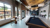 Located at 1415 Iris Dr in Charlotte, NC, this single-floor amenity lounge showcases a compelling industrial-modern aesthetic that blends raw materials with refined furnishings. The space is defined by soaring ceilings adorned with reclaimed wood paneling and exposed ductwork, creating a striking contrast between warmth and utilitarian edge. Polished concrete floors run throughout, tying the open layout together cohesively.

The game area serves as a central attraction, equipped with a billiards table, a full-length shuffleboard table, and a ping-pong table, all positioned beneath pendant drum lighting. Multiple wall-mounted flat-screen TVs add an entertainment focal point along one wall. Large glass garage-style doors and floor-to-ceiling windows flood the space with natural light while framing views of the surrounding neighborhood.

A comfortable lounge area features rich cognac leather sofas and armchairs arranged around low tables, accented by lush potted plants and exposed concrete columns. High-top bistro tables near the entrance provide additional casual seating. The overall ambiance balances an energetic, social atmosphere with relaxed, inviting pockets — making it a well-rounded communal gathering space.