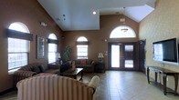 This single-story space features a spacious, open lounge area defined by its dramatic vaulted ceilings that draw the eye upward and contribute to an airy, expansive atmosphere. The warm brown-toned walls provide a cozy backdrop throughout the room, complemented by light tile flooring that reflects natural light and enhances the sense of openness.

Arched windows are a defining architectural element, appearing on multiple walls and flooding the interior with natural light. The grand double entry doors, framed by a prominent arched transom window above, serve as a striking focal point and further reinforce the space's elegant character. A pendant light fixture hangs from the vaulted ceiling, adding a decorative touch near the entrance.

The seating area is comfortably furnished with striped upholstered sofas and armchairs arranged around a dark wood coffee table, creating an inviting setting for socializing or relaxing. Table lamps on side tables provide warm ambient lighting, while decorative accents, including a tall potted plant and small sculptures, add personality to the space. Overall, the room balances architectural character with functional, comfortable furnishings.