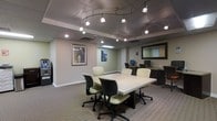 This single-floor business center is thoughtfully designed to provide a functional and professional environment for work and collaboration. The space is anchored by a large, rectangular conference table surrounded by ergonomic cushioned chairs, making it well-suited for meetings or group work sessions. An eye-catching track lighting system spans the ceiling, offering directed, adjustable illumination throughout the room, complemented by recessed lighting for a balanced and well-lit atmosphere. The flooring features neutral-toned carpet that contributes to a quiet, focused work environment. Along one wall, a dedicated workstation area houses desktop computers, providing individual workspace for tasks requiring technology access. A large framed mirror on the wall adds depth to the space, while carefully selected wall art, including a framed abstract piece and a portrait artwork, lend character and visual interest. The muted, neutral color palette of the walls and furnishings creates a calm, professional ambiance. A self-service kiosk or equipment station is also present near the entrance, adding to the space's utility. Overall, the business center is designed with both functionality and comfort in mind, offering a versatile setting for productivity.