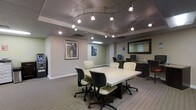This single-floor business center is thoughtfully designed to provide a functional and professional environment for work and collaboration. The space is anchored by a large, rectangular conference table surrounded by ergonomic cushioned chairs, making it well-suited for meetings or group work sessions. An eye-catching track lighting system spans the ceiling, offering directed, adjustable illumination throughout the room, complemented by recessed lighting for a balanced and well-lit atmosphere. The flooring features neutral-toned carpet that contributes to a quiet, focused work environment. Along one wall, a dedicated workstation area houses desktop computers, providing individual workspace for tasks requiring technology access. A large framed mirror on the wall adds depth to the space, while carefully selected wall art, including a framed abstract piece and a portrait artwork, lend character and visual interest. The muted, neutral color palette of the walls and furnishings creates a calm, professional ambiance. A self-service kiosk or equipment station is also present near the entrance, adding to the space's utility. Overall, the business center is designed with both functionality and comfort in mind, offering a versatile setting for productivity.