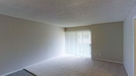This single-floor space offers a clean, functional layout designed for comfortable everyday living. The main living area is carpeted in neutral tones and benefits from generous natural light streaming through a large sliding glass door fitted with vertical blinds, creating a bright and open atmosphere. The textured popcorn ceiling adds a classic touch overhead, while the neutral wall tones provide a versatile backdrop throughout.

A hallway connects the living area to the rest of the unit, featuring durable wood-look plank flooring that contrasts with the carpeted living space. Along the hallway, a set of built-in open shelving units offers practical storage and display space, adding a functional architectural detail to the transition area. The change in flooring material between spaces helps define distinct zones within the layout.

Overall, the space presents a straightforward, well-maintained interior with a neutral color palette that lends itself easily to personalization. The combination of carpeted living areas, hard-surface flooring in transitional spaces, and ample natural light contributes to a functional and inviting environment throughout this single-story home.