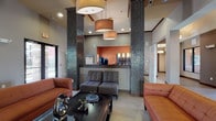 Located in Louisville, Kentucky, the Hurstbourne Estate clubhouse is a thoughtfully designed community facility that blends contemporary style with comfortable, functional spaces. The grand entrance lobby sets the tone with soaring ceilings, striking mosaic tile columns, and bold pendant lighting that casts a warm glow over leather seating and a central reception area accented by rich terracotta tones. The main lounge features a sleek linear fireplace with a mounted flat-screen TV above, complemented by tufted rust-colored sofas, upholstered accent chairs, and large windows that invite natural light throughout. Deep teal walls in the secondary lounge create a cozy, sophisticated atmosphere, furnished with plush seating and an additional wall-mounted TV. The fully equipped kitchen boasts dark cabinetry, granite countertops, a mosaic tile backsplash, stainless steel appliances, and vibrant terracotta walls, ideal for community gatherings. A dedicated business center offers workstations along a granite desk surface beneath pendant lighting and framed artwork. The dining area provides casual seating under elegant pendant lights, while wide-plank tile flooring and high ceilings with crown molding tie the entire space together with a polished, inviting ambiance.