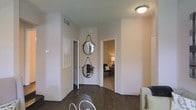 This single-story space presents a cohesive and well-appointed interior defined by neutral white walls and rich dark hardwood flooring that runs throughout. The hallway area serves as a central connector between rooms, offering a glimpse into an adjoining bedroom furnished with a lamp, seating, and warm ambient lighting that creates an inviting atmosphere. Two circular rope-hung mirrors are mounted vertically on the wall, adding a decorative focal point that brings character and visual interest to the otherwise understated corridor. A flush-mount ceiling light provides clean, even illumination. Doors along the hallway lead to additional spaces, suggesting a practical and efficient floor plan. The open sightlines between areas contribute to a sense of flow and continuity throughout the home. The overall aesthetic is modern yet approachable, combining simple architectural lines with thoughtful decorative touches. The consistent use of neutral tones across walls and trim creates a calm, versatile backdrop well-suited to a variety of furnishing styles. The space balances functionality with subtle design details that elevate everyday living.