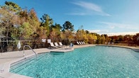 This single-level community amenity space is designed to support an active lifestyle, featuring two distinct areas: a fully equipped fitness center and an expansive outdoor pool complex. The fitness center is bright and welcoming, with neutral-toned walls, crown molding, and multiple windows fitted with white blinds that allow generous natural light to fill the room. The space houses a comprehensive selection of equipment, including treadmills, an elliptical trainer, a stationary bike, and a cable strength machine, as well as a dedicated weight bench and leg extension machine. A wall-mounted flat-screen television and a medicine ball rack with multiple weighted balls round out the amenities, while durable rubber flooring provides a safe, practical workout surface throughout. Adjacent to the fitness center, the outdoor pool area is equally impressive. The large, rectangular pool features sparkling blue water and is surrounded by a spacious concrete deck appointed with lounge chairs for relaxing in the sun. A covered patio with ceiling fans and comfortable outdoor seating offers a shaded retreat, perfect for unwinding. The entire pool area is enclosed by a black wrought-iron fence, with a lush backdrop of mature trees providing privacy and a natural, scenic setting.