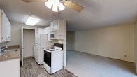 This single-floor space offers an open-concept layout where the kitchen flows directly into the living area, creating a practical and connected environment. The kitchen is equipped with white cabinetry that extends to the ceiling, providing ample storage. Essential appliances include a white refrigerator, an electric range with a black oven, and an over-the-range microwave. The kitchen features hardwood-style flooring, adding a warm contrast to the neutral tones throughout the space. The adjoining living area is finished with carpeting, creating a clear visual distinction between the two zones. A textured ceiling runs throughout, and a ceiling fan with a light fixture is centrally positioned in the living area, offering both ventilation and lighting. The overall color palette is neutral, with beige and white tones dominating the walls, cabinetry, and appliances, contributing to a clean and understated ambiance. The open layout between the kitchen and living space makes the unit feel functional and easy to navigate.