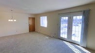 This single-floor space offers a clean, functional layout designed for comfortable everyday living. The main living area is finished with plush wall-to-wall carpeting in a neutral tone, providing a warm and inviting atmosphere throughout. Smooth, light-colored walls create a bright and airy feel, complemented by an abundance of natural light streaming in through a standard window and a set of elegant French doors. The French doors serve as a standout architectural feature, featuring multi-pane glass panels that open to an outdoor area, allowing views of the exterior and flooding the interior with sunlight. A solid wood entry door provides a traditional and sturdy point of entry to the space. The flooring vents suggest a central forced-air heating and cooling system, ensuring year-round climate comfort. The overall color palette—featuring soft beige walls, white trim, and light carpeting—creates a cohesive and understated aesthetic that lends itself well to a variety of furnishing styles. The open layout of the living area allows for flexible furniture arrangements, making the space highly adaptable. The combination of natural light, neutral finishes, and practical design elements makes this a straightforward and welcoming single-level space.