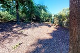 This outdoor space is a dedicated dog park enclosed by chain-link fencing, offering a natural and shaded environment for pets to roam freely. The ground is covered with wood chip mulch throughout, providing a comfortable and low-maintenance surface underfoot. Mature trees are distributed across the space, casting generous shade and contributing to a calm, park-like atmosphere under open skies.

The perimeter is fully secured with chain-link fencing, ensuring safe containment for pets. A metal bench is positioned near the entrance, providing a convenient resting spot for owners. The wooded surroundings, made up of dense shrubs and established trees, create a sense of natural enclosure and separation from the adjacent multi-story residential building visible beyond the fence line.

The layout is open and spacious, allowing ample room for pets to move around freely. The combination of mature tree canopy, wood chip coverage, and secure fencing makes this a functional and well-maintained amenity. Natural light filters through the tree canopy, illuminating the space while the surrounding greenery adds to the overall tranquil and nature-integrated character of the area.