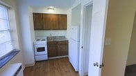This single-floor space features a compact, efficiently laid-out kitchen that makes practical use of its footprint. The kitchen is outfitted with rich, dark wood cabinetry both above and below the counter, providing ample storage. A stainless steel sink is centrally positioned beneath the upper cabinets, complemented by a white electric range and a full-size white refrigerator. The flooring throughout is a warm hardwood-style material that adds a touch of warmth to the space. Neutral white walls keep the area feeling clean and bright, while a flush-mount ceiling light provides overhead illumination. A doorway visible on the right side of the kitchen suggests convenient access to an adjacent room, contributing to the overall functional flow of the layout. The space is straightforward and move-in ready, suited for those seeking a practical, low-maintenance living environment.