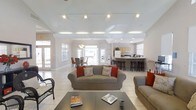 This single-story space showcases a thoughtfully designed open-plan layout that integrates the living, dining, and kitchen areas into one cohesive, flowing environment. The living area is anchored by comfortable upholstered sofas accented with rust-colored throw pillows, arranged around a dark coffee table that grounds the space. Light tile flooring extends throughout, contributing to the airy, clean aesthetic.

Overhead, the ceiling features distinctive exposed beams and a track lighting system, providing both architectural character and flexible illumination. Recessed lighting supplements the natural light that pours in through multiple large windows and French doors, enhancing the bright, welcoming atmosphere.

The dining area is positioned toward the rear left, furnished with a classic table and chairs and illuminated by a pendant light fixture. French doors nearby suggest access to an outdoor area. The kitchen occupies the rear of the space, equipped with light wood cabinetry, a white refrigerator, and a raised breakfast bar with dark bar stools — ideal for casual dining or entertaining. White walls and neutral tones throughout unify the space, creating a clean, inviting environment.