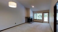 This single-floor space centers around a well-appointed living area that combines comfort and functionality. The room features vaulted ceilings that lend a sense of openness and volume, while neutral-toned walls and light carpet throughout create a clean, versatile backdrop. A corner fireplace with a brick surround and raised hearth serves as a natural focal point, adding warmth and character to the space. Large windows positioned adjacent to a glass-paneled exterior door flood the room with abundant natural light and offer appealing views of mature green trees outside, creating a strong connection to the surrounding landscape. Recessed lighting and a pendant light fixture provide layered illumination options. A floor-level vent indicates a forced-air heating system integrated throughout. The open layout of the living area allows for flexible furniture arrangements, making it well-suited for everyday living and entertaining alike. Overall, the space presents a balanced combination of architectural interest through its vaulted ceilings and fireplace, practical amenities, and a bright, welcoming ambiance enhanced by natural light and outdoor greenery.