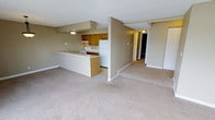 This single-floor residential space offers a functional and welcoming open-concept layout that seamlessly connects the kitchen and living area. The kitchen is equipped with warm honey-toned wood cabinetry, white appliances including a refrigerator, range, and range hood, and features a peninsula counter that serves as both a workspace and a divider between the kitchen and the main living area. The peninsula provides additional counter space and adds to the room's sense of organization and flow.

The living area is generously proportioned, featuring plush, neutral-toned carpeting that extends throughout the space, lending a warm and comfortable atmosphere. The walls are finished in a soft beige tone that complements the carpeting and cabinetry, creating a cohesive and understated color palette. Flush-mounted ceiling lights provide even illumination throughout.

A hallway visible from the main living space leads to additional areas of the unit, hinting at further rooms beyond. A set of bi-fold closet doors is also visible, offering convenient built-in storage. The overall layout is practical and efficient, with clear sightlines across the main living area. The neutral finishes and open arrangement make the space versatile and easy to personalize.