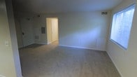 This single-floor space features an open living area finished with wall-to-wall neutral carpeting that provides a clean, understated foundation. The walls are painted in a soft, light tone that contributes to a bright and airy atmosphere throughout the room. Visible HVAC floor vents and ceiling registers indicate a centralized climate control system, ensuring year-round comfort. A doorway in the background leads to an adjacent area, suggesting a straightforward and functional layout. An electrical panel is visible along one wall, offering practical accessibility. White baseboards frame the perimeter of the room, adding a subtle, clean finish to the overall aesthetic. The space is vacant, allowing for flexible furniture arrangements to suit a variety of needs. Overall, the unit presents a modest, move-in-ready environment with a neutral palette well-suited for personalization.