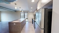 This well-appointed one-bedroom, one-bathroom residence at 1464 E 105th St in Cleveland, Ohio offers a thoughtfully designed single-floor layout with a clean, modern aesthetic. Light wood-tone flooring runs throughout the unit, complementing the neutral warm-white walls and crisp white baseboards that give the space a bright, cohesive feel.

The kitchen is a standout feature, showcasing a large central island with a granite-style countertop and bold blue cabinetry base, pendant lighting overhead, and a full suite of stainless steel appliances including a refrigerator, electric range, microwave, and dishwasher. Two-tone cabinetry — pairing gray lower cabinets with lighter upper cabinets — lines the perimeter walls, and a large window admits generous natural light with views of the surrounding neighborhood.

The open living area is spacious and filled with natural light from floor-to-ceiling windows that frame outdoor neighborhood views, creating an airy and inviting atmosphere. The hallway, featuring exposed ductwork overhead, connects the main living spaces to the private bedroom, which is comfortably sized and includes a window with roller shade.

The bathroom is finished with large-format tile flooring, a warm wood-toned vanity with white countertop, a full mirror, and a glass-enclosed shower/tub combination. The entry area includes built-in panel storage, adding practical functionality to the unit's overall efficient design.