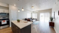 Located at 79 N 1020 W in American Fork, Utah, this single-floor residence offers a thoughtfully designed open-concept layout that seamlessly connects the kitchen, dining, and living areas. The kitchen is a functional centerpiece, showcasing white upper cabinetry, dark lower cabinetry, a light quartz countertop island with seating, stainless steel appliances including a refrigerator, range, and microwave, and elegant pendant lighting overhead. Wood-look flooring flows throughout the main living areas, complementing the neutral color palette and creating a cohesive, modern aesthetic. The living room is bright and welcoming, with multiple windows dressed in white blinds that invite abundant natural light, and a sliding glass door that provides outdoor access. A chandelier adds a refined touch to the space. The bedroom features plush carpeting, a generously sized window, and direct access to a private en-suite bathroom equipped with a vanity and ample storage. An additional full bathroom includes a tub-shower combination, toilet, and wood-look flooring. Throughout the unit, clean lines, textured ceilings, and well-placed recessed and overhead lighting contribute to a comfortable and contemporary living environment suited for everyday functionality.