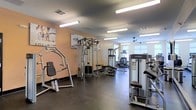 This single-floor fitness center offers a spacious and functional workout environment designed to accommodate a wide range of exercise needs. The facility is outfitted with a robust selection of Life Fitness strength training machines, including cable and pulley systems, weight benches, and various resistance stations, providing ample options for both targeted muscle training and full-body conditioning. Cardio equipment is also present, complementing the strength-focused layout.

The interior features a neutral color palette with warm peach-toned walls, accented by motivational fitness posters that contribute to an energizing atmosphere. Durable dark flooring spans the workout area, offering both practicality and a clean aesthetic. Overhead fluorescent lighting ensures the space is well-lit throughout, while a ceiling fan toward the rear of the room aids in air circulation. Natural light filters in through windows at the far end of the facility, adding brightness and a sense of openness to the environment. The layout is thoughtfully organized, allowing for comfortable movement between equipment stations and supporting an efficient, focused workout experience.