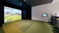 Located at 3111 Sunset Boulevard in Flower Mound, TX, this thoughtfully designed indoor golf simulator suite offers a compelling blend of sport and sophisticated hospitality. The space is anchored by a dedicated simulator bay finished with realistic turf flooring and a large projection screen, delivering an immersive golf experience. Dark wall paneling in the simulator zone creates a dramatic contrast against the bright, open lounge area that adjoins it.

The lounge is finished with wide-plank light oak hardwood flooring and features a striking floor-to-ceiling wainscoting accent wall in soft white, lending a refined, architectural character to the space. Comfortable seating, including plush armchairs and a modern sofa with warm tones, is arranged thoughtfully to accommodate spectators and guests. Sculptural accent tables with brass-toned bases add an elegant design touch throughout.

A built-in bar area with light wood cabinetry, a beverage refrigerator, and a clean white countertop provides a hospitality-ready setup for entertaining. Curated wall art, including framed golf-themed prints and a vibrant painting, adds personality and visual interest. Recessed lighting throughout ensures the entire suite is well-lit and inviting. The overall ambiance is modern and upscale, balancing athletic function with a welcoming, lounge-like atmosphere.