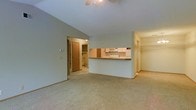 This single-floor residence offers a thoughtfully arranged open-concept layout that maximizes functionality and flow. The main living area is spacious and welcoming, featuring soft wall-to-wall carpeting that adds warmth and comfort underfoot. A ceiling fan is centrally positioned overhead, providing both light and air circulation throughout the space.

The kitchen is defined by a half-wall pass-through counter with a wood-toned laminate surface, creating a natural separation between the cooking and living areas while maintaining an open feel. Light-colored upper cabinetry lines the kitchen walls, complemented by a microwave and countertop appliances that hint at a practical, efficient layout.

A doorway visible from the main living area leads to what appears to be a utility or laundry closet, complete with shelving and washer/dryer hookups — a convenient and practical inclusion. An additional hallway on the opposite side of the space suggests access to further rooms, offering a sense of depth and connectivity throughout the floor plan.

The neutral color palette — soft beige walls, cream-toned ceilings, and warm carpeting — creates a clean, cohesive aesthetic that is easy to personalize. Overall, this single-floor space combines practicality with a comfortable, open atmosphere well-suited for everyday living.