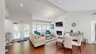 Located at 7303 Bryn Athyn Way in Raleigh, NC, this well-appointed single-floor space offers a thoughtfully designed layout comprising a living room, kitchen, and hallway. The living room is the centerpiece of the space, showcasing soaring vaulted ceilings that lend an airy, open feel. A white-mantled fireplace serves as a striking focal point, complemented by rich hardwood flooring and a stylish globe chandelier. Banks of large windows fitted with horizontal blinds line the walls, flooding the room with abundant natural light while framing views of the surrounding greenery. The dining area flows seamlessly from the living room, featuring the same hardwood flooring and a generous wall of windows that connect the interior to the outdoors. A glass-topped dining table with upholstered chairs anchors this inviting space. The kitchen is clean and functional, outfitted with crisp white cabinetry, polished dark granite countertops, and a breakfast bar with seating. Stainless steel appliances, including a built-in microwave, add a modern touch, while a window over the counter brings in natural light. The neutral gray-white color palette throughout the space creates a cohesive, contemporary ambiance that feels both comfortable and refined.
