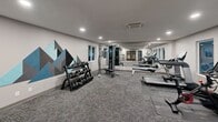 Located at 7654 40th St W in University Place, WA, this well-appointed fitness center offers a comprehensive workout environment designed for both convenience and performance. The single-floor facility features recessed lighting throughout, a textured ceiling, and a combination of durable wood-look plank flooring and commercial-grade carpet tiles that define distinct activity zones.

The main workout area is generously proportioned and lined with floor-to-ceiling mirrors along one wall, visually expanding the space while allowing users to monitor their form. A standout decorative mural of geometric mountain peaks in shades of blue and gray adds a striking visual focal point. Equipment includes a multi-station cable machine with a dedicated bench, a tiered dumbbell rack stocked with a wide range of weights, treadmills, an elliptical, and a stationary spin bike — all positioned to maximize space and flow.

Windows along the perimeter bring in natural light and offer views of the adjacent outdoor pool area, creating an energizing atmosphere. A dedicated locker nook features built-in shelving, coat hooks, and a warm wood bench for storage and changing. A touchless water bottle filling station is conveniently mounted nearby. Climate control is provided via a ceiling-mounted HVAC unit, ensuring year-round comfort. A keypad-secured entry door provides controlled access to the facility.