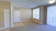 Located at 7111 N Blue Angel Pkwy in Pensacola, Florida, this single-floor residence offers a comfortable and functional layout with a clean, neutral aesthetic throughout. The living area is carpeted and generously sized, bathed in natural light from a large sliding glass door fitted with vertical blinds that span nearly the full wall, creating a bright and airy atmosphere. A ceiling fan with a light fixture is centrally mounted, adding both comfort and practical illumination to the space. Adjacent to the living area, a hallway leads toward the bedrooms, maintaining a straightforward and accessible floor plan. The entry area transitions from tile flooring to carpet, providing a defined threshold between the entryway and the main living space. Track lighting is installed near the front entry, offering adjustable, directed lighting in that zone. A window with horizontal blinds allows additional natural light into the entryway. The kitchen, visible from the living area, features dark countertops and dark cabinetry, providing a subtle contrast to the warm beige tones of the walls. Overall, the space presents a neutral color palette with beige walls, light carpet, and white trim, lending it a versatile and move-in-ready feel suitable for a variety of furnishing styles.