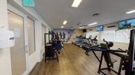 This single-floor fitness center offers a comprehensive workout environment designed to support a wide range of exercise routines. The space is laid out across an open, spacious floor plan finished with warm hardwood-style flooring throughout, creating a clean and inviting atmosphere.

The cardio zone is well-stocked with multiple treadmills, elliptical machines, and stationary bikes from brands including True and Fatigue, arranged in rows to maximize floor space. Wall-mounted flat-screen televisions are positioned throughout the room, providing entertainment during workouts. A dedicated alcove features additional treadmills alongside large windows that offer outdoor views and bring in natural light.

The strength training area includes Cybex cable machines, a leg press, dumbbell racks, barbells, weight plates, and a variety of benches and resistance machines, catering to both beginners and experienced lifters. A wall of mirrors along one side enhances the sense of space and allows users to monitor their form.

Ceiling fans contribute to air circulation, complementing the ventilation system overhead. Motivational large-format wall murals — including a dynamic running silhouette graphic and an ocean wave scene — add an energizing visual element to the environment. Flush-panel ceiling lighting ensures the space is brightly and evenly illuminated throughout.