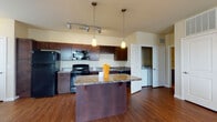 This 1-bedroom, 1-bathroom residence offers a thoughtfully designed open-concept layout that seamlessly connects the kitchen, dining, and living areas. The kitchen is a standout feature, showcasing rich dark wood cabinetry, granite countertops, a full suite of black appliances including a refrigerator, electric range, built-in microwave, and dishwasher, as well as a wine rack cabinet and a central island with pendant lighting overhead. Track lighting adds additional ambiance and functionality to the space. Hardwood-style flooring flows throughout the main living areas, complementing the warm, neutral color palette of the walls. The living room is bright and airy, benefiting from multiple windows that invite natural light and offer outdoor views, along with a ceiling fan for added comfort. A private entrance door from the living area adds convenience. The bedroom is carpeted for comfort, features its own ceiling fan and a generously sized window, and provides direct access to a hallway leading to a closet and the full bathroom. The bathroom includes a tiled tub-and-shower combination, hardwood-style flooring, and dark vanity cabinetry with granite countertops. A dedicated in-unit laundry area is also included, adding everyday practicality to this well-equipped residence.