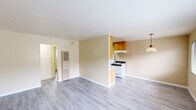 This single-floor unit offers a thoughtfully arranged layout comprising a living room, bedroom, kitchen, bathroom, and hallway. The interior is finished with light gray hardwood-style flooring throughout, creating a cohesive and clean aesthetic across all rooms. Walls are painted in a soft, warm cream tone that contributes to a bright and neutral ambiance.

The living area is open and spacious relative to the overall footprint, with a wall-mounted HVAC unit and a pendant light fixture visible near the kitchen opening. The kitchen is designed in a galley configuration, featuring warm wood cabinetry, a dual-basin stainless steel sink, speckled countertops, a white gas range, and a full-size refrigerator — providing ample functionality within a compact space.

The bedroom receives natural light through a window fitted with horizontal blinds, and includes an wall-unit air conditioner, offering comfortable climate control. The bathroom is equipped with a single-sink vanity with under-cabinet storage, a large wall mirror with overhead lighting, and a tub-shower combination with white tile surround and a small window for ventilation. A hallway connects the rooms, adding a sense of structure to the overall layout.