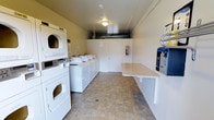 This single-floor shared laundry facility is designed with practicality and efficiency in mind. The room is laid out to maximize usability, featuring a row of top-loading washing machines along one wall and a stacked washer-dryer unit positioned prominently near the entrance. The machines are uniformly white, contributing to the clean, orderly appearance of the space.

A wall-mounted folding table runs along one side of the room, providing a convenient surface for sorting and folding laundry. Adjacent to it, a vending machine offers easy access to laundry supplies. A fire extinguisher is visibly mounted on the wall, reflecting attention to safety standards.

The flooring consists of neutral-toned tile, which is both durable and easy to maintain. The walls are painted in a light, muted tone, keeping the space feeling open and tidy. Overhead lighting provides adequate illumination throughout. Exposed ductwork is visible near the ceiling at the back of the room, adding an industrial touch. A utility door at the rear provides access to mechanical areas. Overall, the space is well-maintained and thoughtfully organized for shared use.