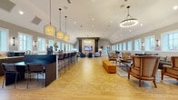 Located at 142 Patrick Murphy Dr in Morganton, NC, this single-story community clubhouse offers a thoughtfully designed, open-plan gathering space ideal for socializing and events. The interior is defined by soaring vaulted ceilings and wide-plank hardwood flooring featuring an eye-catching geometric parquet pattern at the center, adding a distinctive architectural touch underfoot.

The space is anchored by a dramatic feature wall housing a large flat-screen display, flanked by dark stone columns and a textured wood panel backdrop. A circular chandelier provides warm ambient lighting over the lounge area, which is furnished with contemporary sofas, accent chairs, and coffee tables arranged to encourage conversation. Stylish pendant lights—including cage-style and hoop fixtures—are suspended throughout, contributing to the room's modern aesthetic.

A long, dark-topped bar-height counter with black metal stools runs along one side of the room, complemented by a fully equipped kitchenette featuring white subway tile backsplash, marble-look countertops, dark cabinetry, and stainless steel appliances. Rows of white-trimmed windows line the perimeter walls, flooding the interior with natural light and views of the surrounding community. Additional bistro-style seating nooks are available for smaller gatherings, making this a versatile and welcoming amenity space.