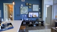 This single-floor space showcases a thoughtfully designed interior that blends modern aesthetics with industrial influences. The living area immediately draws attention with its striking deep blue accent wall, which serves as a bold backdrop for a carefully arranged gallery wall featuring an eclectic mix of framed artwork in varying sizes and styles. A wall-mounted flat-screen television anchors the display, flanked by open metal media consoles that provide functional storage while reinforcing the space's industrial character. Pendant lighting with an exposed cage-style fixture adds warmth and a touch of urban flair overhead. The wood-look flooring grounds the space with natural texture, complementing the cool tones of the walls. A light-colored surface, likely a kitchen counter or bar, is visible in the foreground along with a round-topped stool, suggesting a casual dining or bar area that flows seamlessly into the living space. Decorative accents — including ceramic vases, small sculptures, and stacked books — are thoughtfully placed throughout, adding personality and depth. A soft area rug defines the seating zone, contributing to the overall warmth and livability of this well-curated, open-concept space.