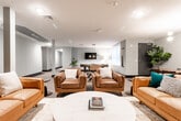 Located on E Dickerson Street in Dover, NJ, this single-floor amenity space is designed to offer residents a well-rounded social and recreational experience. The layout flows across several distinct zones, each thoughtfully curated with modern finishes and cohesive design elements throughout.

The main lounge area serves as a central gathering hub, furnished with plush caramel leather sofas arranged around a round white coffee table. A large-scale mural accent wall provides a dramatic backdrop for the wall-mounted flat-screen TV, while warm lighting and lush potted plants add texture and life to the space.

Adjacent to the lounge, the game room is a standout feature, equipped with a full-size billiards table, a foosball table, and a wall-mounted television. Industrial-style pendant lighting and a window with natural light create an energetic yet comfortable atmosphere for recreation.

A private dining area offers a more intimate setting, centered around a large wooden dining table surrounded by black Windsor-style chairs. An elegant brass and globe chandelier illuminates the space, while a full-length mirror and flanking fiddle-leaf fig trees lend a refined, sophisticated touch.

Connecting these areas is a spacious corridor with neutral carpeting, structural columns, and clean sage-green walls, reinforcing the space's contemporary aesthetic throughout.