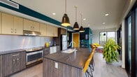 Located at 2155 E Lark St in Springfield, Missouri, this single-floor space showcases a sleek, contemporary design defined by bold teal accent walls, dark wood cabinetry, and large-format tile flooring. The open-concept layout seamlessly connects a fully equipped kitchen to a comfortable lounge area, creating a cohesive and functional environment.

The kitchen is a standout feature, outfitted with dark wood-grain lower cabinetry paired with lighter upper cabinets, dark stone countertops, a stainless steel appliance suite including a range, dishwasher, and refrigerator, and a metallic backsplash. A central island with a built-in sink and bar seating provides additional workspace and a casual dining option. Matte black pendant lights hang overhead, adding a modern industrial touch.

The adjacent lounge area features floor-to-ceiling windows and a glass wall with black-framed double doors leading to an outdoor patio, flooding the space with natural light and offering open views of the surrounding landscape. The living zone is furnished with vibrant yellow accent chairs arranged around a round white coffee table, creating an inviting gathering spot.

Throughout, recessed lighting, clean architectural lines, and the striking teal color palette establish a cohesive, modern aesthetic with an energetic yet welcoming ambiance.